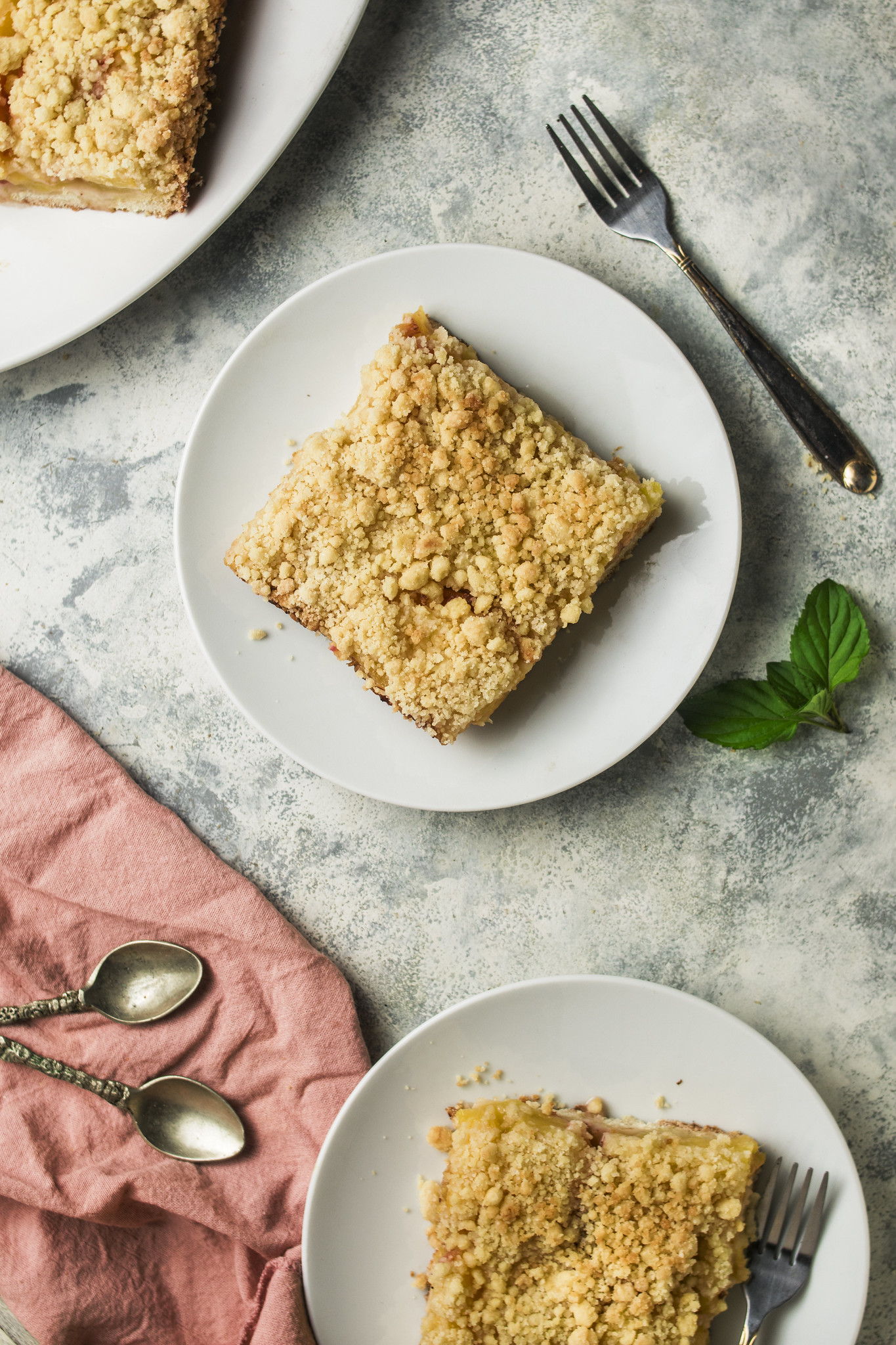 Overhead view of sbriciolata di pesche served on white plates, with crumbly peach cake slices, vintage cutlery and a pink cloth on a softly textured surface.